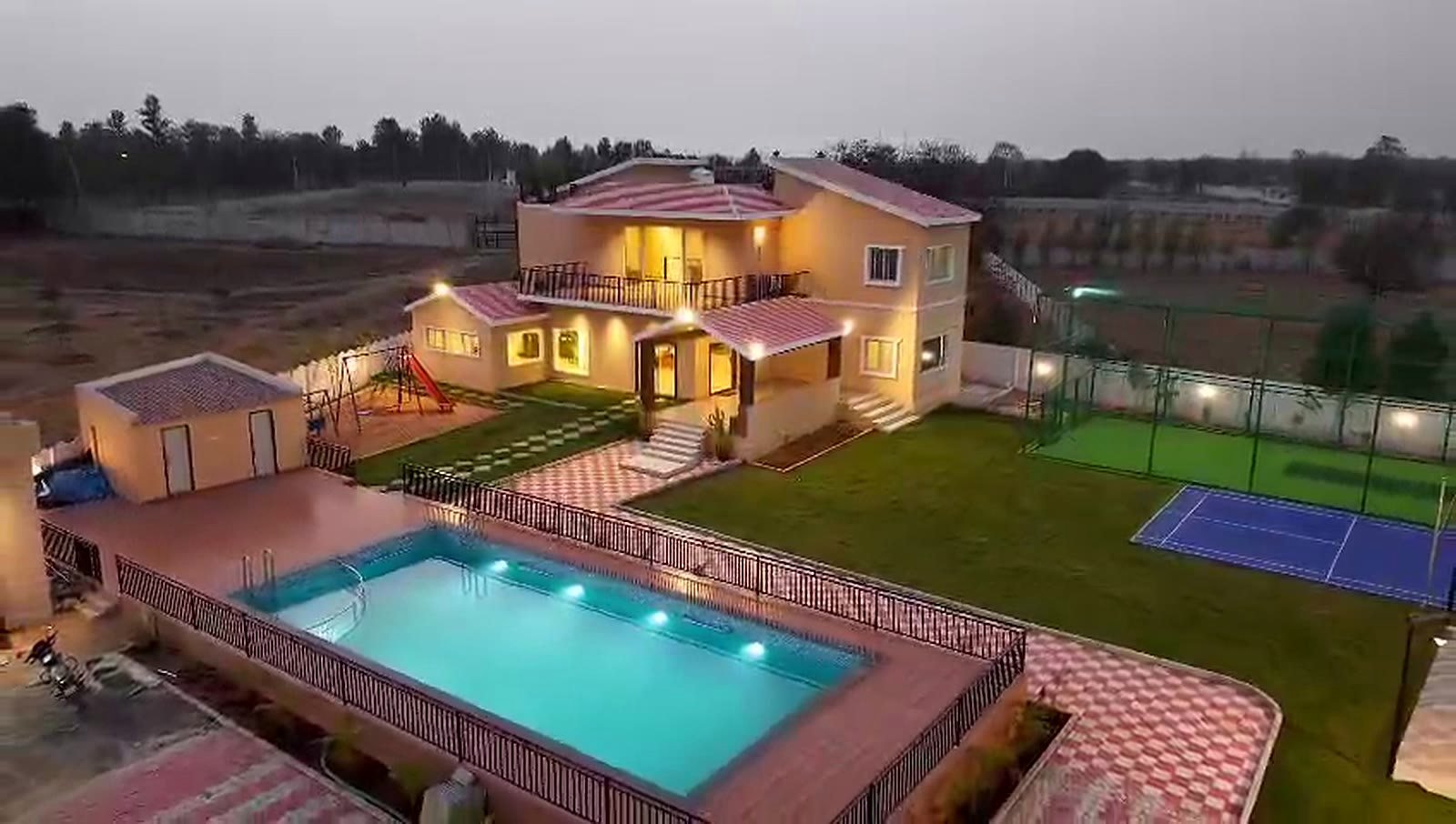 An elevated evening view over the lit pool, the lawn and the sports courts.